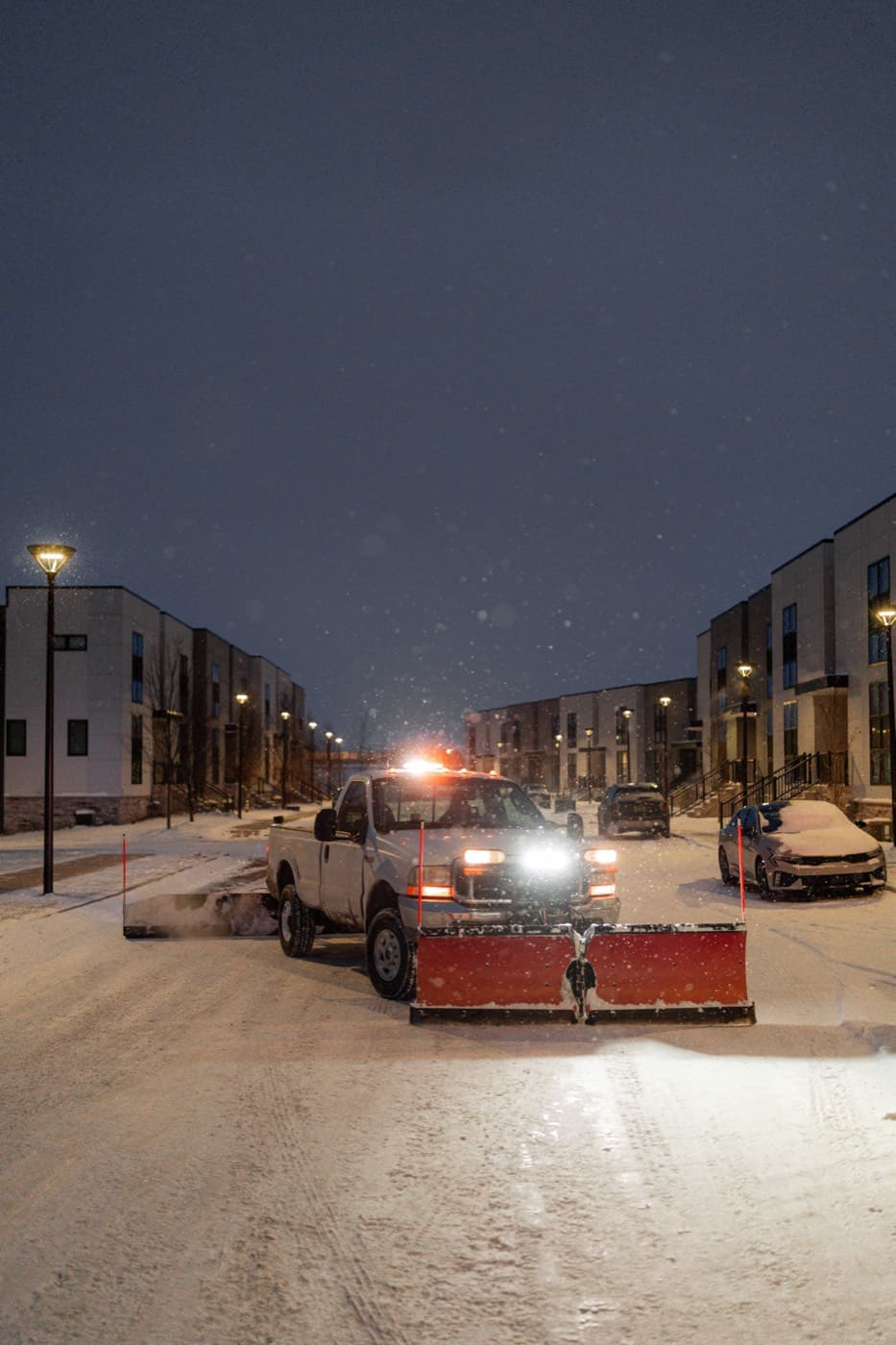 Rodas Premier plow truck with its blade down, actively plowing a snow-covered street between townhomes during a Michigan snowfall